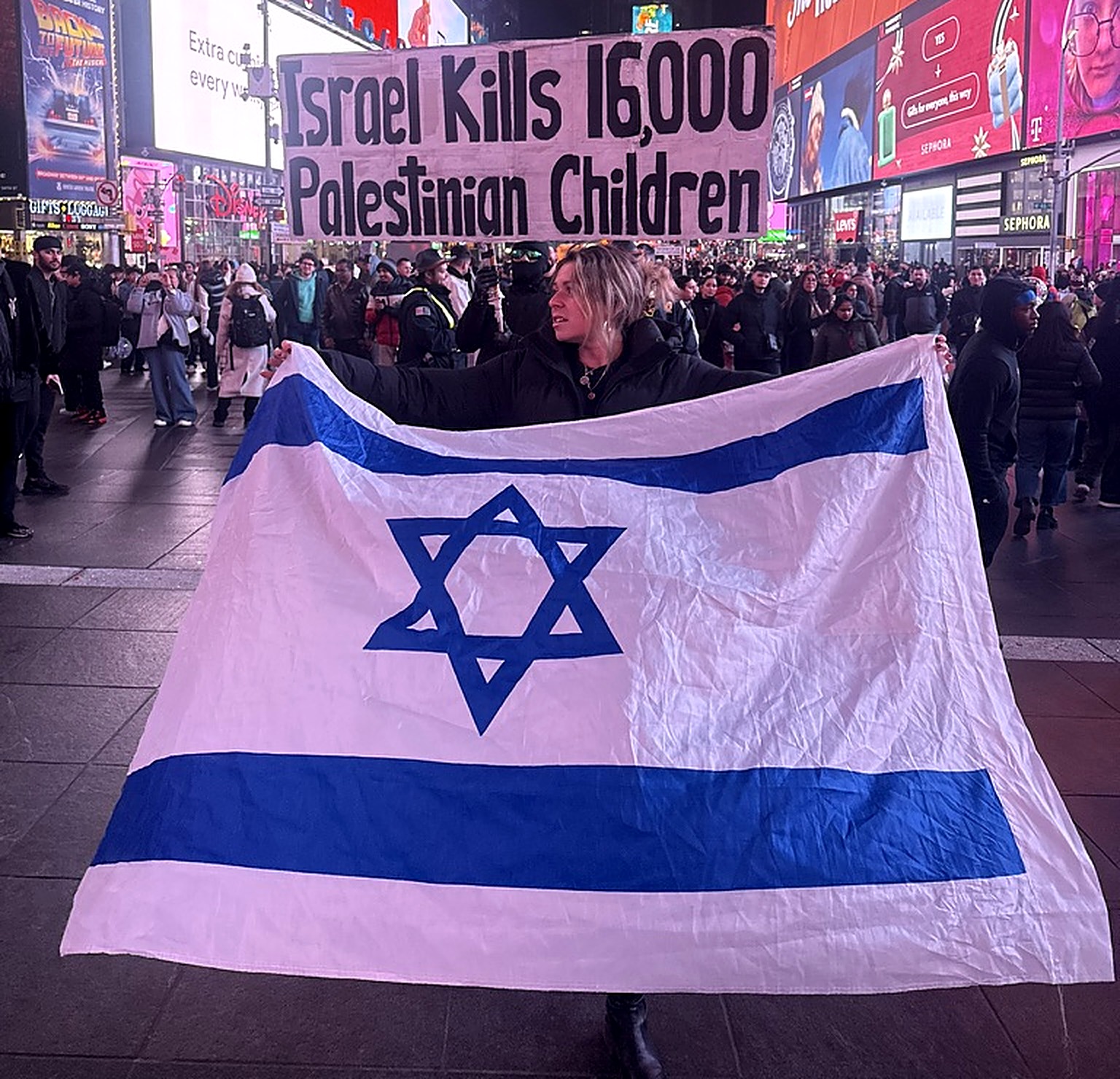 Chana holding a large Israeli flag in Times Square at night, facing an anti-Israel protest sign